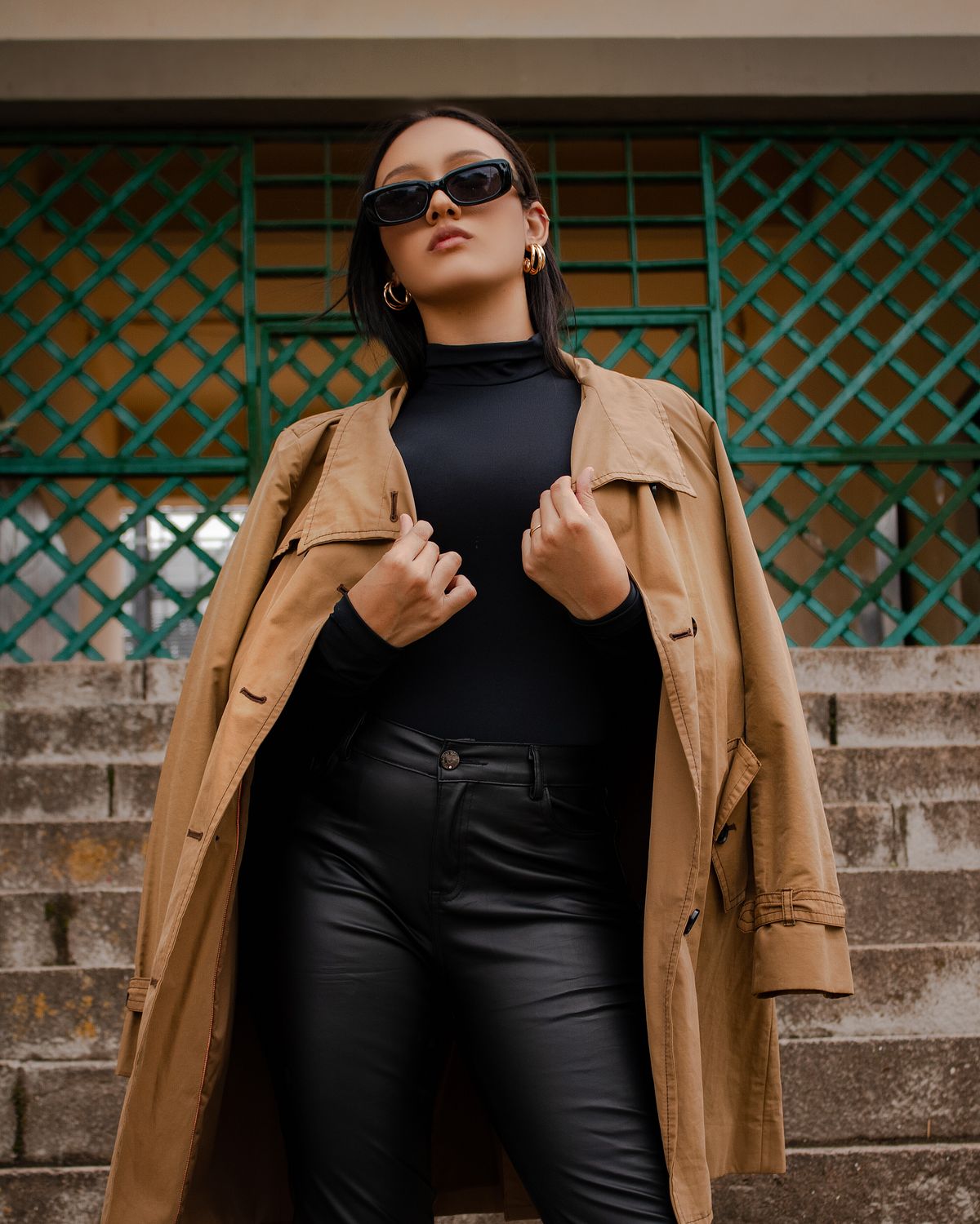 Fashionable woman with sunglasses poses confidently on city stairs, wearing a trendy trench coat.