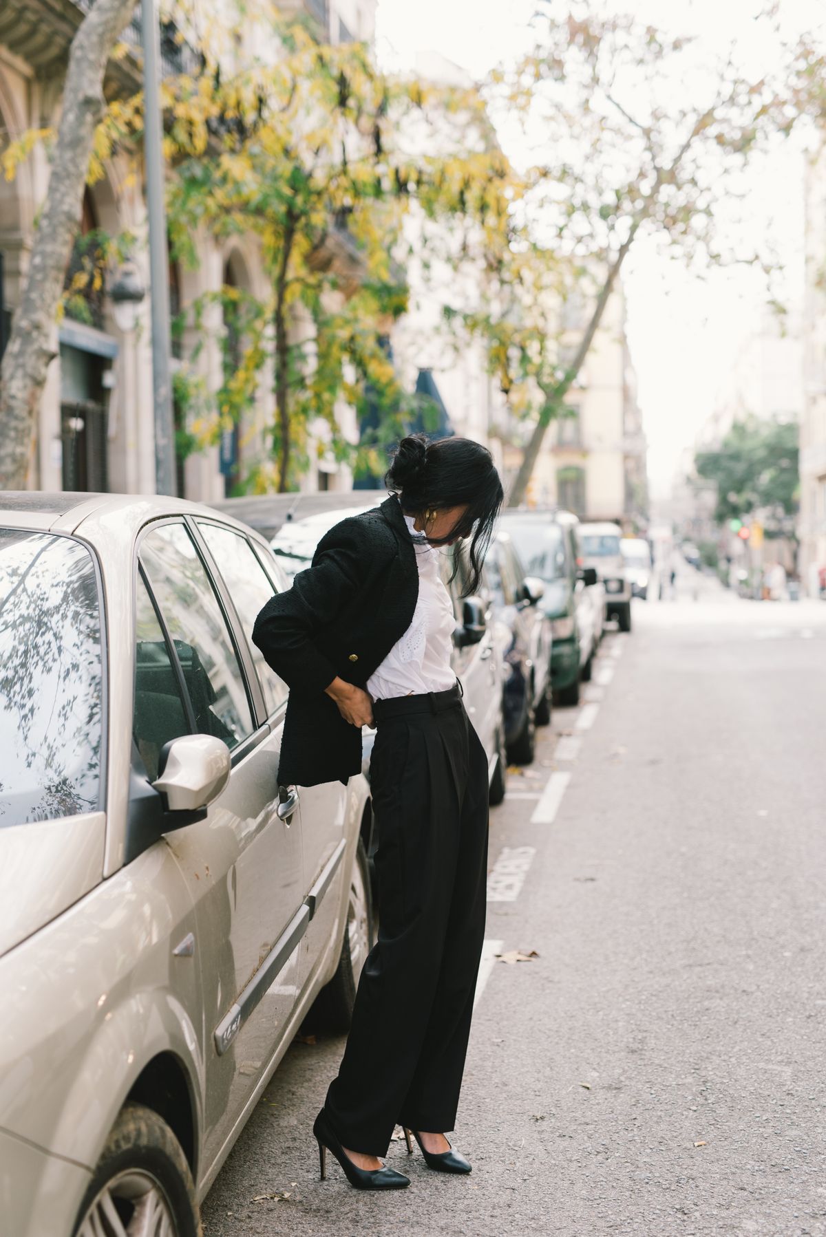 Fashionable woman in a black coat and heels standing by parked cars on a tree-lined street.